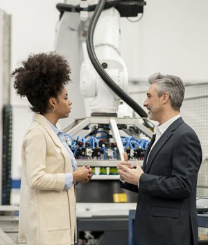 Two professionals in business attire stand in front of a large robotic arm and industrial machinery, engaged in conversation.