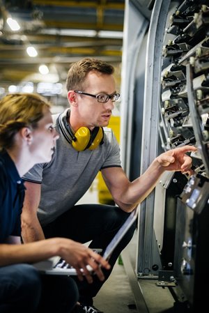 Two individuals, one wearing yellow ear defenders, are working together in a factory setting, examining and interacting with a complex piece of industrial machinery while one holds a laptop.