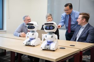 Two white robots with blue lights sit on a wooden table in front of a group of people observing them.